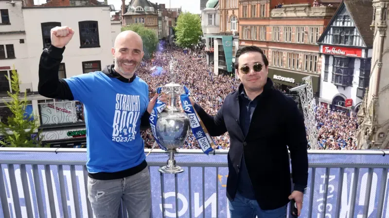 Leicester City manager Enzo Maresca and owner Top Srivaddhanaprabha with the EFL Championship trophy. Behind them are fans watching on celebrating the title.