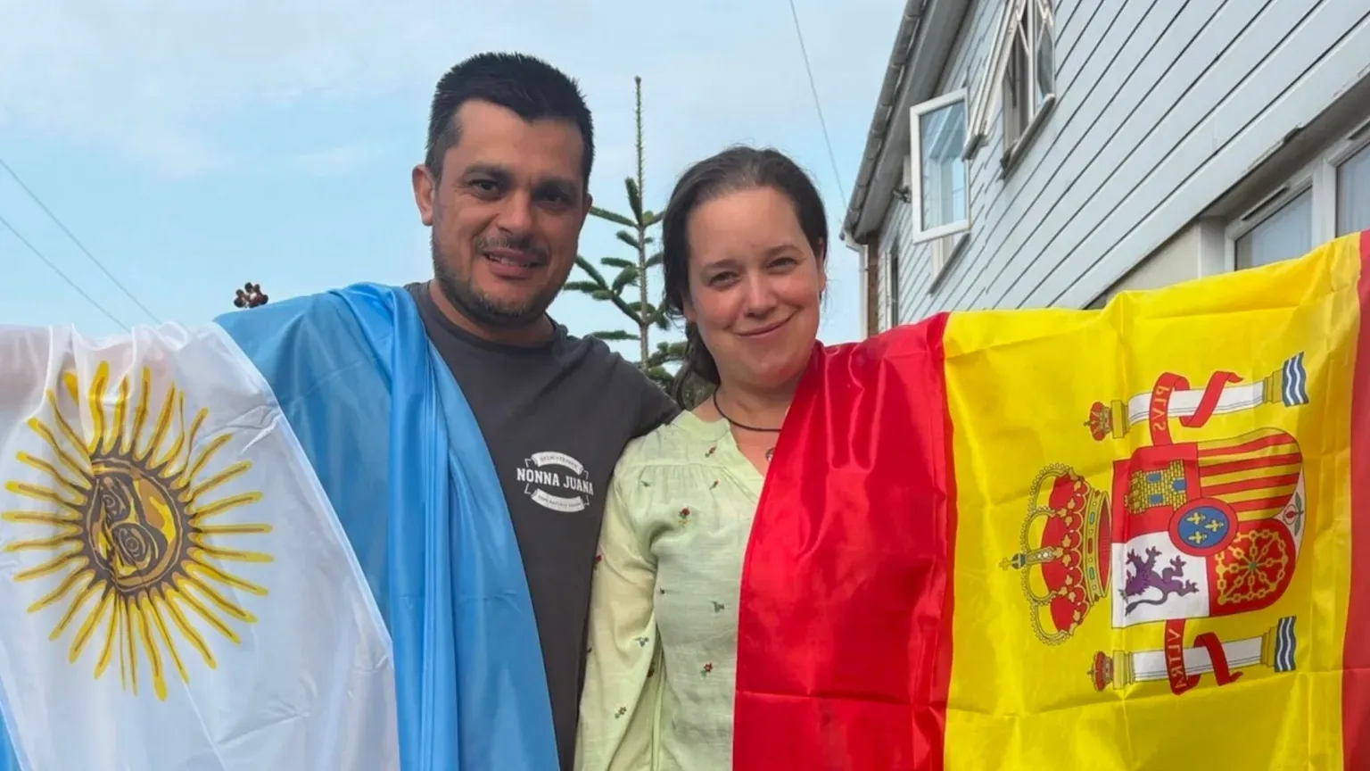 Herraiz family A man and a woman stand side by side holding their country's flags. The man has short dark hair and a beard and is wearing a grey top. He is holding the blue and white flag of Argentina, which is draped over his shoulder. Maria has shoulder-length hair and is wearing a yellow patterned shirt. She is holding the red and yellow flag of Spain.