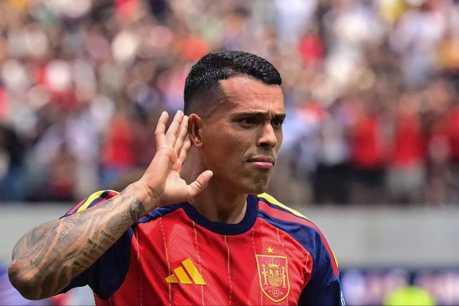 Pedro Porro cups his right hand to his right ear in celebration after scoring the second for Spain. He is wearing their red home shirt and is before a blurred background of a crowd at SoFi Stadium