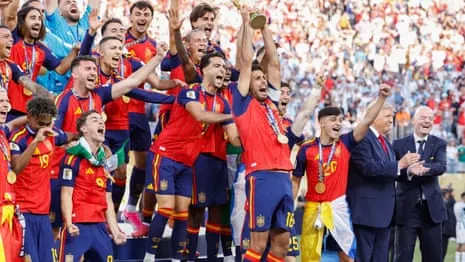 Spain celebrate with the World Cup trophy on a podium, arms raised, with officials applauding.
