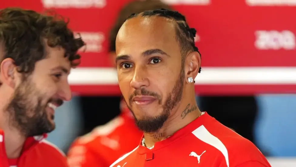 Lewis Hamilton is wearing a red and white top, has short black hair in tight cornrows and has a trimmed black beard. He is starting to smile.
