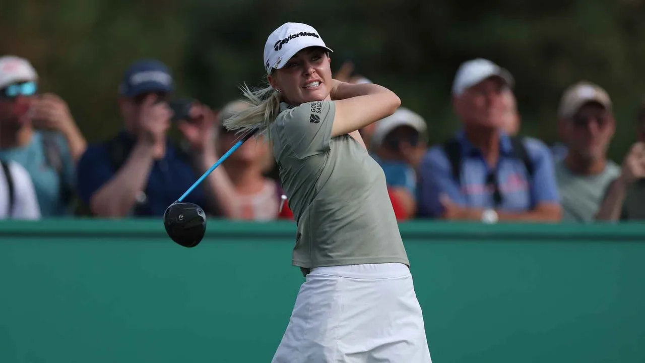 A female golfer swings a driver during a game, wearing a cap and light-colored shirt with a white skirt. Spectators with cameras watch from behind a green barrier.